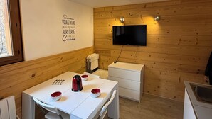 Une cuisine compacte avec une table blanche, des tasses rouges et blanches, une bouilloire et un téléviseur fixé à un mur en bois.