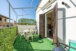Un balcon avec une table et des chaises, un gazon synthétique vert, un mur blanc et une porte à volets marron.