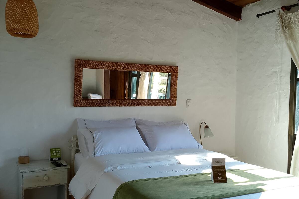 Un dormitorio con techo de madera, una cama con ropa de cama blanca, un edredón verde, una mesita de noche con una lámpara y un espejo en la pared.