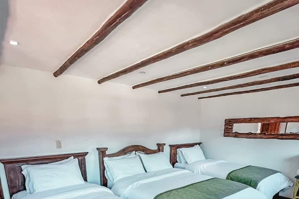 Habitación de hotel con tres camas de madera, ropa de cama blanca y estructura de vigas de madera en el techo.
