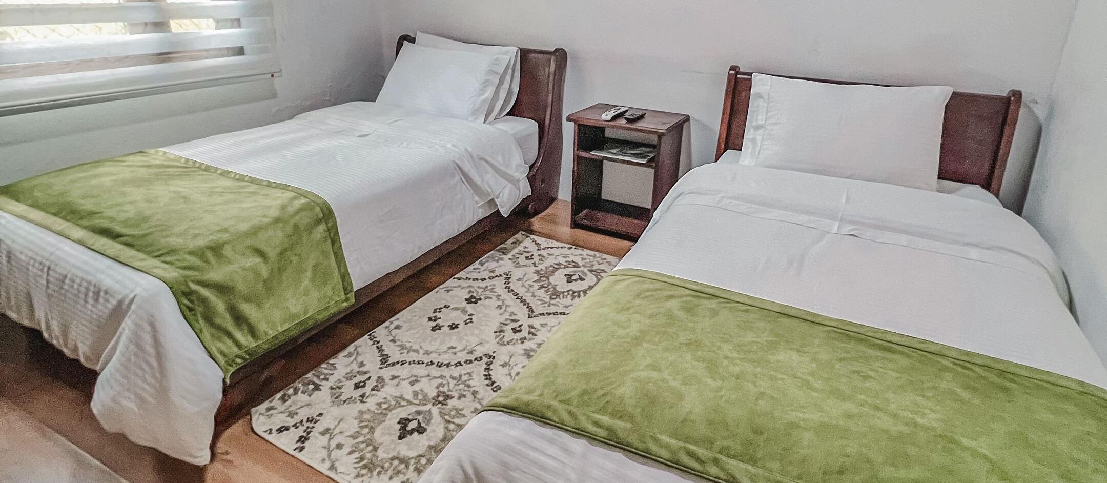 Une chambre avec deux lits, chacun recouvert d’une couverture verte, une petite table de chevet avec un téléphone et un tapis à motifs.