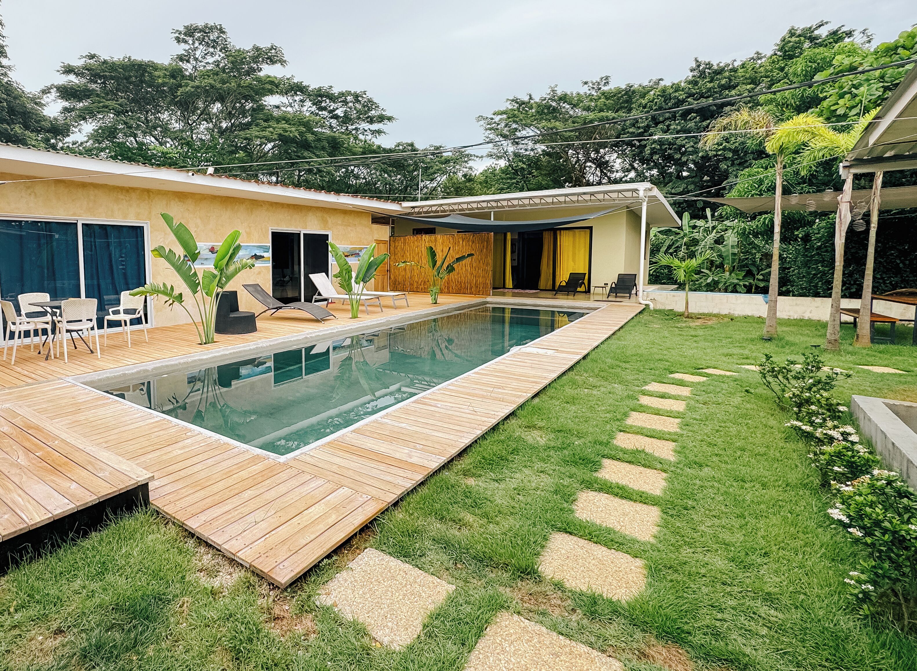 A swimming pool with a wooden deck, surrounded by a grassy area with stepping stones, and a building with a covered patio.