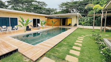 A swimming pool with a wooden deck, surrounded by a grassy area with stepping stones, and a building with a covered patio.