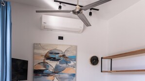 Un salon moderne avec un canapé gris, une table basse en bois, un ventilateur de plafond, un climatiseur fixé au mur, un tableau, une horloge et un téléviseur accroché au mur.