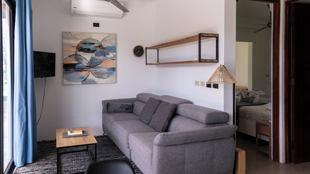 A modern living room with a grey sofa, a small wooden table, a ceiling fan, a wall-mounted clock, and a painting. - Modern Hideaway with Pool –  Near Sámara Location! (Sámara)