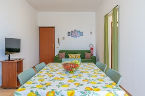 Un coin repas avec une table dressée pour quatre personnes, un téléviseur posé sur un meuble, et un canapé vert en arrière-plan.