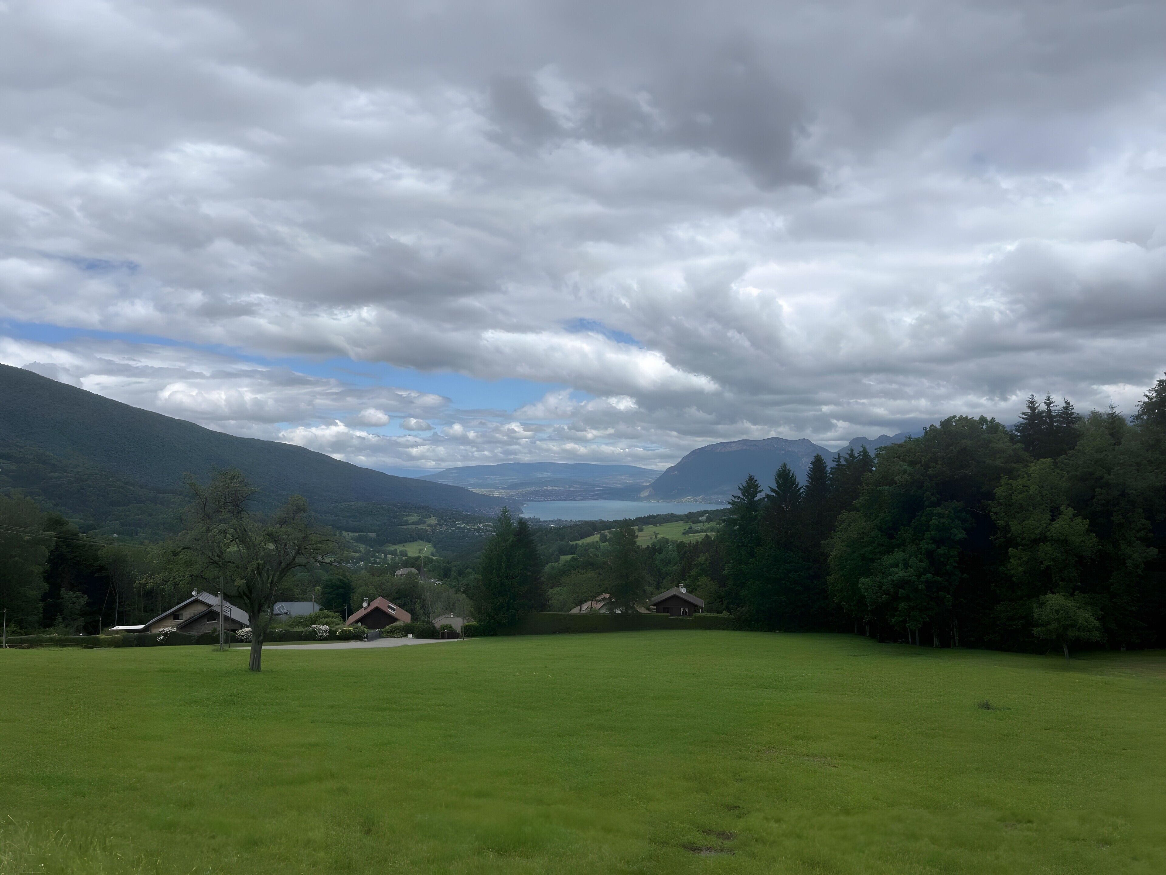 Ländliche Landschaft mit Häusern, einer großen grünen Wiese und Bergen in der Ferne unter einem bewölkten Himmel.