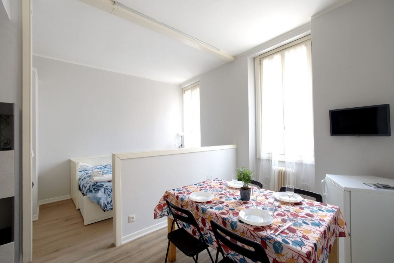 Una stanza piccola e luminosa con un letto, un tavolo da pranzo apparecchiato per due persone, una televisione e una finestra con tende.