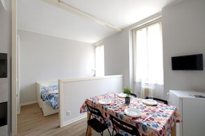 Ein kleiner, hell erleuchteter Raum mit einem Bett, einem Esstisch für zwei Personen, einem Fernseher und einem Fenster mit Vorhängen.