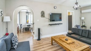 A modern living room with a sofa, a coffee table, a TV mounted on the wall, and a dining area in the background. - 4BR Pitt Suburbs Gem (Pittsburgh)