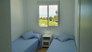 A room with two single beds, a small bedside table, and a window with a view of a grassy area and the sea. - Vista al Mar con terraza-Apartamento Costa Ballena (chipiona)