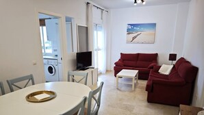A compact living space with a dining table, two red sofas, a TV, and a painting of a beach scene. - Vista al Mar con terraza-Apartamento Costa Ballena (chipiona)
