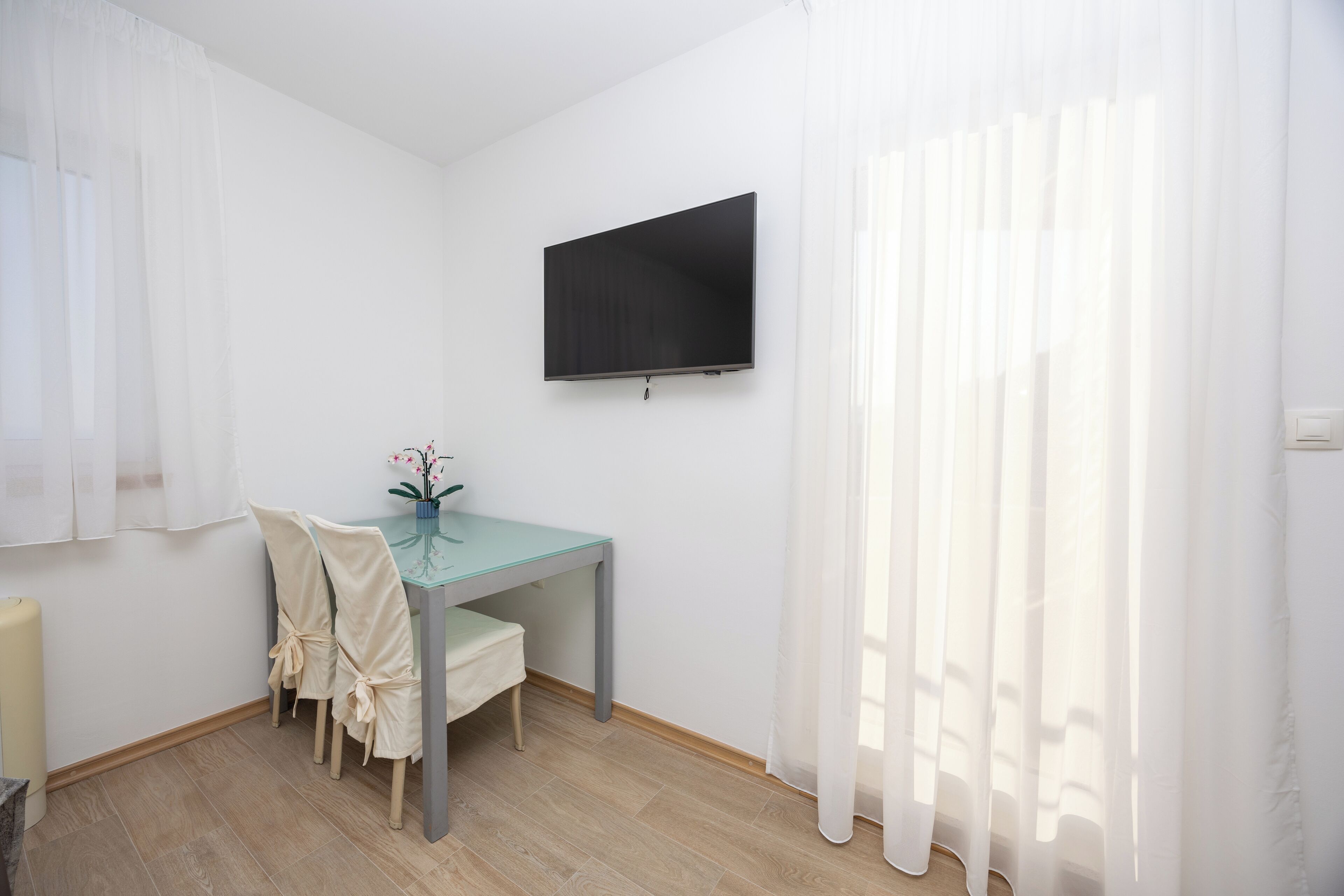 A small, bright room with a wooden floor, a table with chairs, a flat-screen TV mounted on the wall, and sheer white curtains.