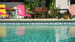 Un área junto a la piscina con diferentes sillas y sombrillas, un borde de piscina con azulejos y vegetación al fondo.