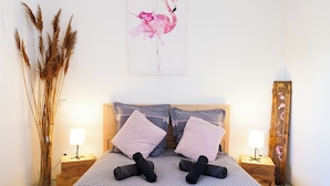 Una cama bien hecha con colcha de lunares, dos almohadas y un cabecero de madera. Hay dos mesitas de noche con lámparas, un jarrón con plantas secas y una obra de arte en la pared con un flamenco.