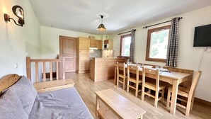 Een knus woongedeelte met een bank, eettafel, stoelen, een keukenhoek en een aan de muur bevestigde televisie.