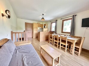 Kompakti oleskelutila, jossa on sohva, ruokapöytä, tuolit, keittiöalue ja seinälle asennettu televisio.