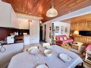 A cozy living room with a dining table set for four, a kitchen area, a red sofa, and a television.