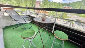 A balcony with green artificial turf, two bar stools, a round table with a red polka-dotted bowl, and a view of a mountainous area with buildings. - 50m from ski slopes, 4th floor, view swimming pool/mountain, ski locker, 45m², Vars (Vars)