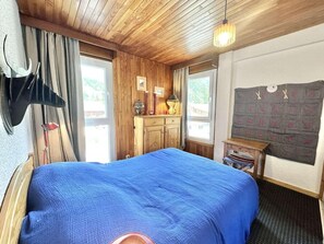 A bedroom with a large bed, wooden ceiling, a small wooden dresser, a nightstand, and a window with curtains.