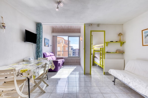 A room with a bed, a sofa, a table, a TV, and a window with a view of buildings.