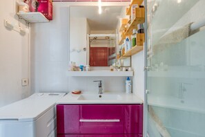 Un baño con un lavabo blanco, un espejo, cabina de ducha de vidrio y un mueble rosa.