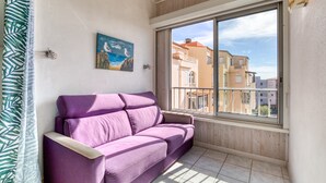 A room with a purple sofa, a painting of seagulls, a window with a view of buildings, and a wooden ceiling. - Apartment 'Sur Le Port' with Sea View and Private Garden (Agde)