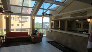 A hotel lobby with a red sofa, a wicker chair, a reception desk, and a large window with a view of buildings and trees. - N.Myrtle Beach Oceanfront 2BR, July 20-27 Shorecrest Vacation Villas I resort (North Myrtle Beach)