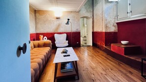Un espacio habitable compacto con un sofá, un sillón blanco, una mesita con un mando a distancia y una zona de cocina con armarios y un cuenco sobre una encimera.