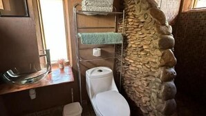 Une petite salle de bain avec un mur en pierre, des toilettes blanches, une étagère en métal avec des serviettes et un petit lavabo.