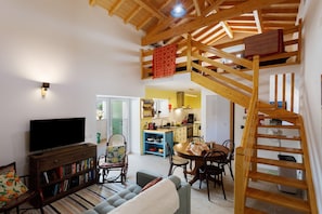 Un espacio habitable tipo loft con una televisión, estanterías, un sofá, una zona de comedor y una cocina al fondo.