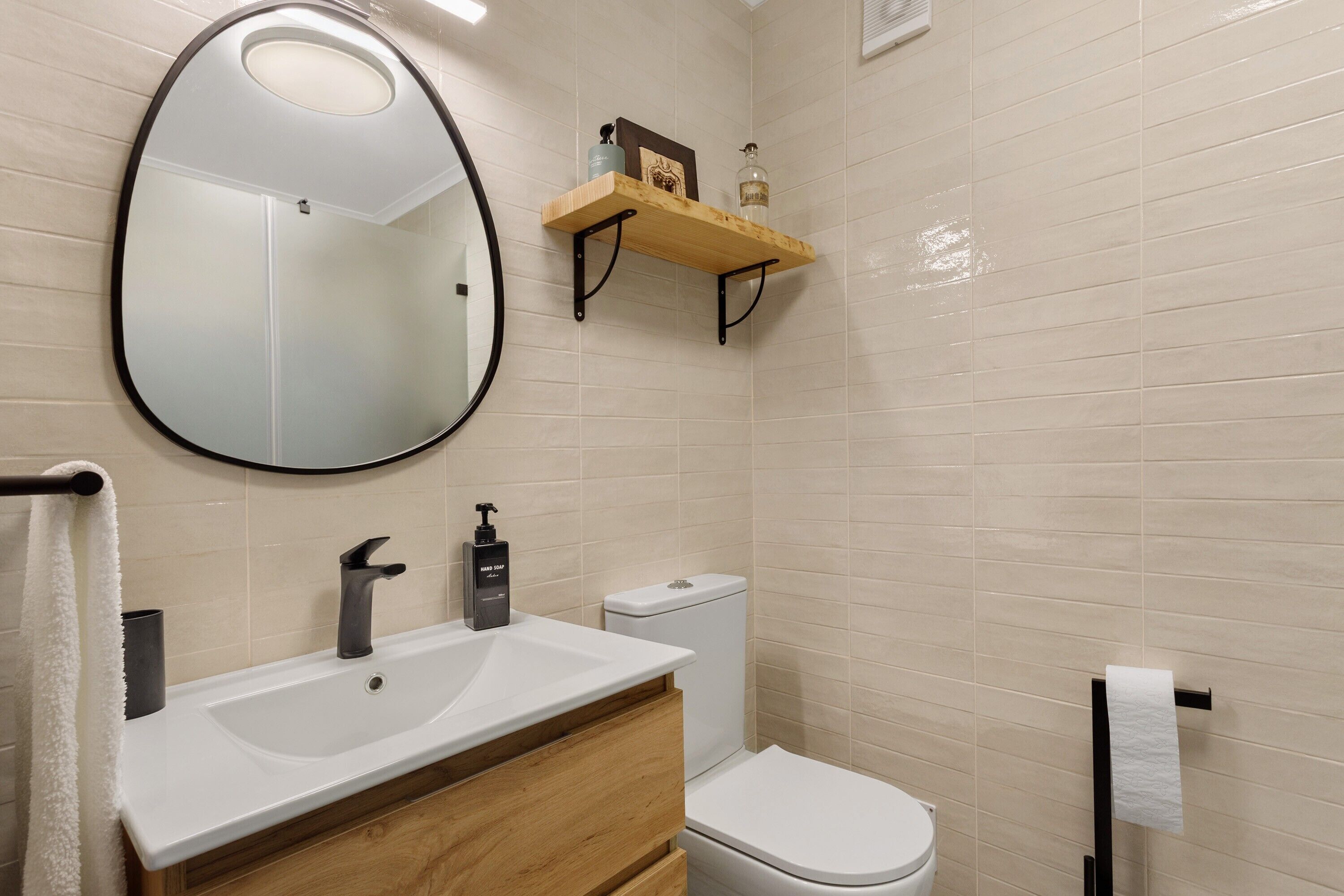 Une salle de bain avec un lavabo blanc, un robinet noir, un meuble-lavabo en bois, des toilettes blanches et un miroir avec un cadre noir.