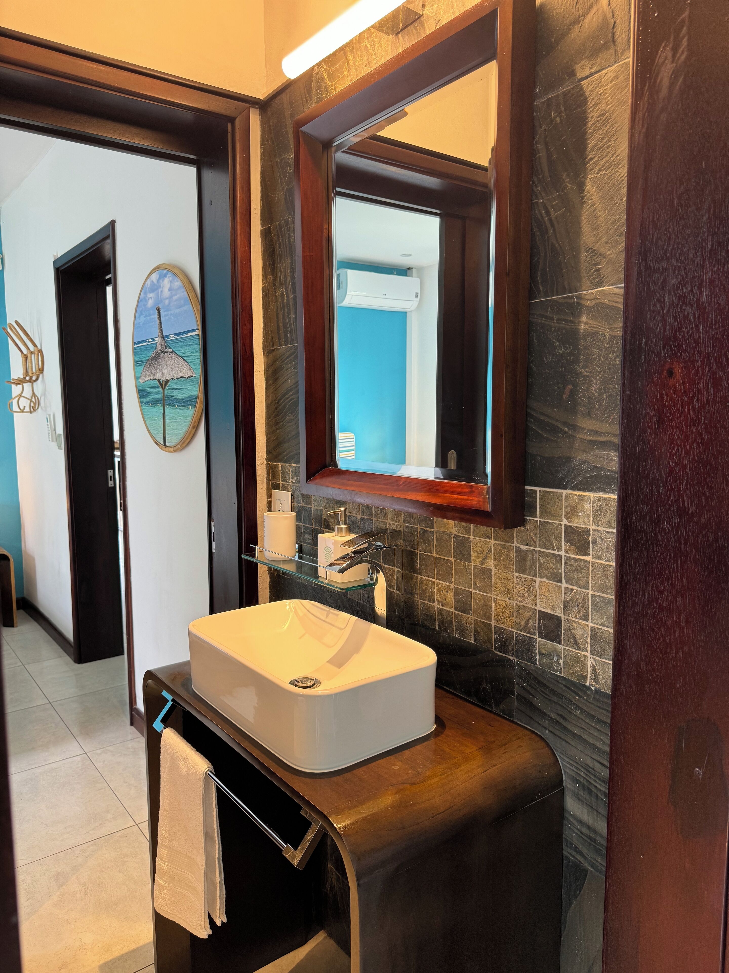 A bathroom with a white sink, a glass shelf with toiletries, a marble backsplash, and a door leading to another room.