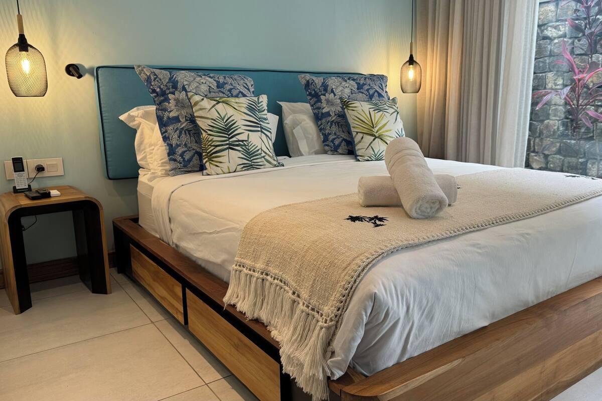 Een bed met houten frame, een blauw hoofdbord en meerdere kussens. Er staat een nachtkastje met een telefoon en een stopcontact.