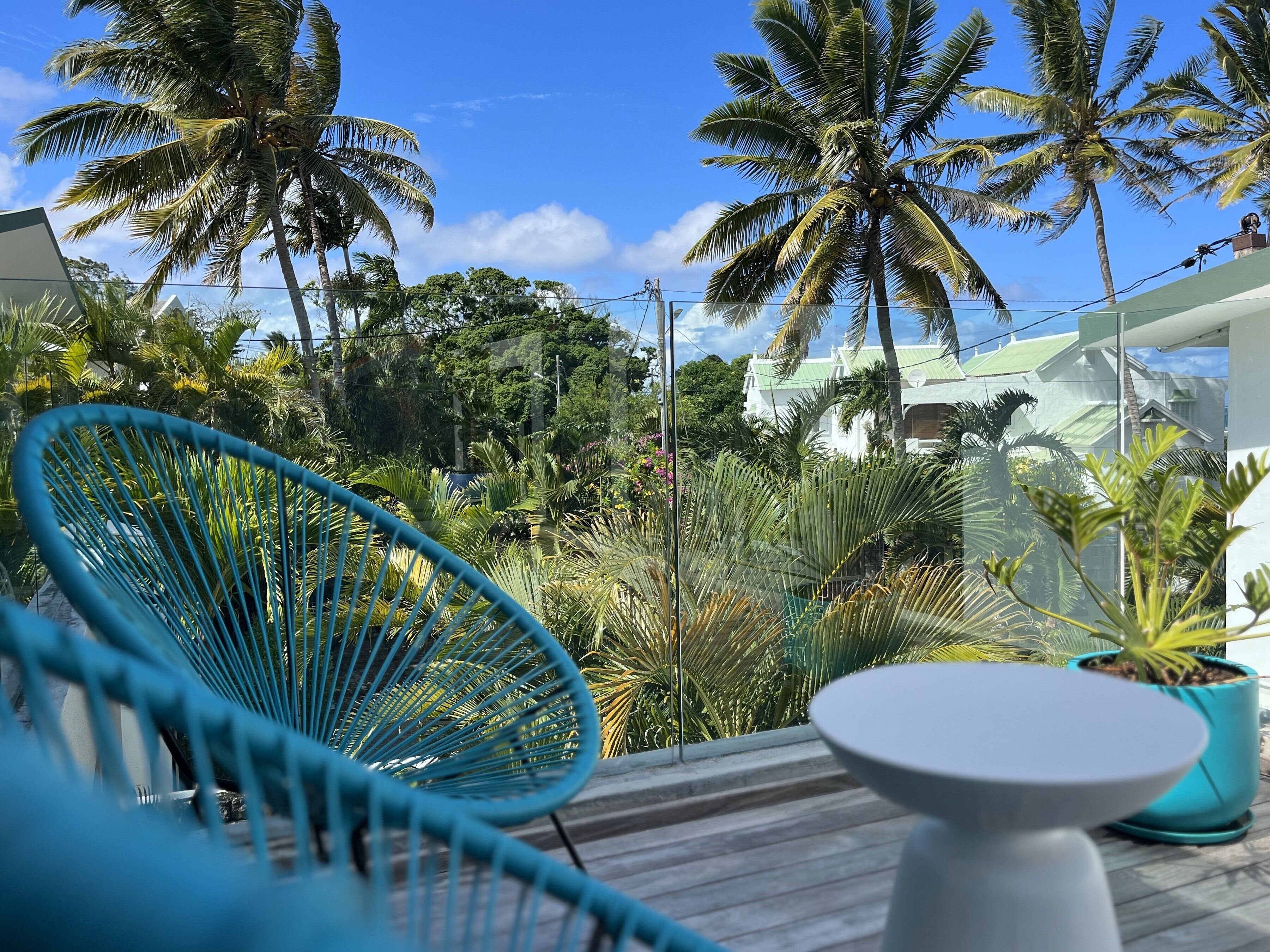 A balcony with a blue chair, a small table, and a view of palm trees and a building in the background.