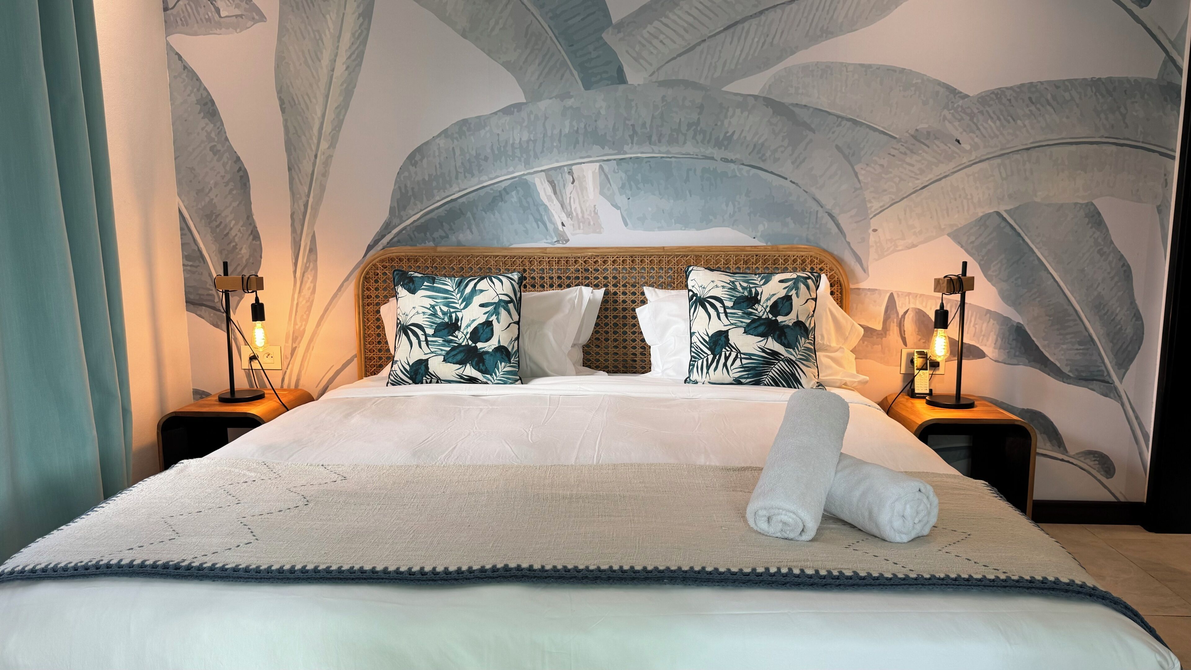 A large bed with a textured headboard, two nightstands with lamps, and a tropical leaf-patterned wall mural.
