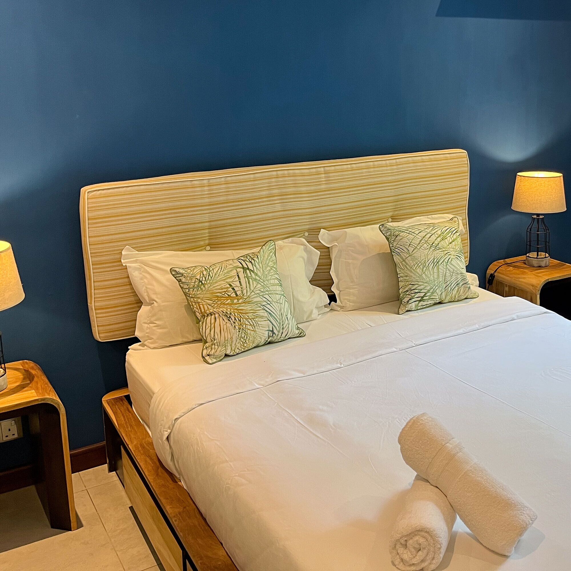 Een net opgemaakt bed met witte lakens en twee decoratieve kussens, een houten hoofdbord en een bedlampje op een houten nachtkastje.