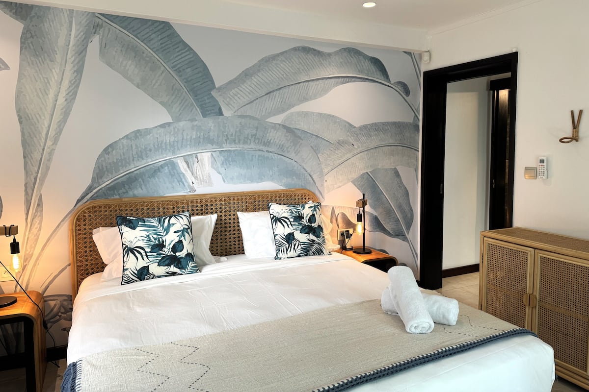 Een net opgemaakt bed met een witte dekbedovertrek en bloemenkussens, een houten hoofdbord en een wanddecoratie van grote bladeren.