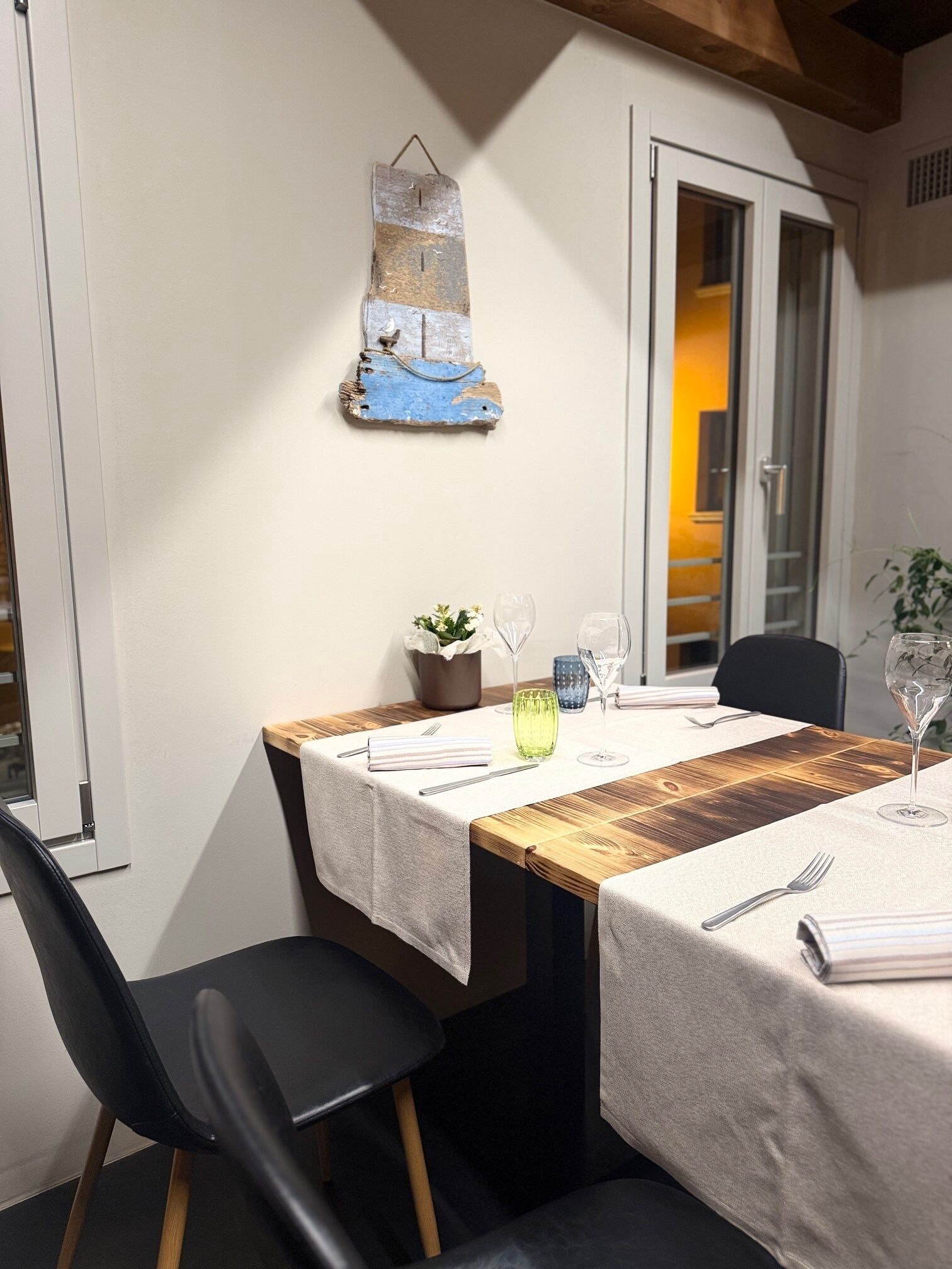 Un coin repas avec une table en bois dressée pour deux personnes, une plante en pot et un élément décoratif fixé au mur.