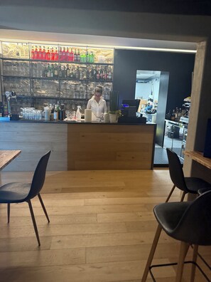 Una zona de bar con mostrador de madera, estantes con botellas y dos sillas negras.