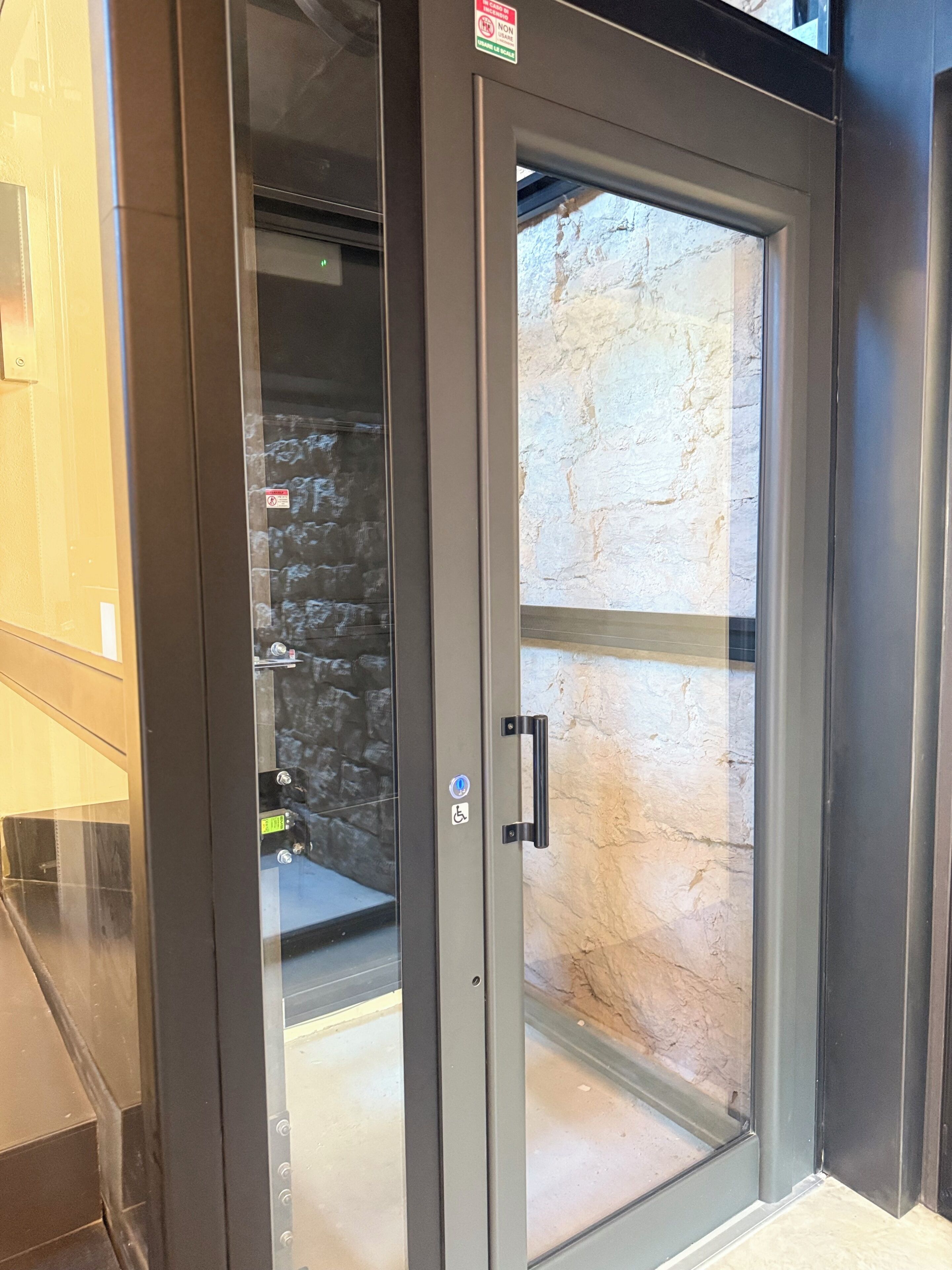 Een moderne lift met glazen deur, een rolstoeltoegankelijkheidsbordje en een zichtbare stenen muur door de deur.