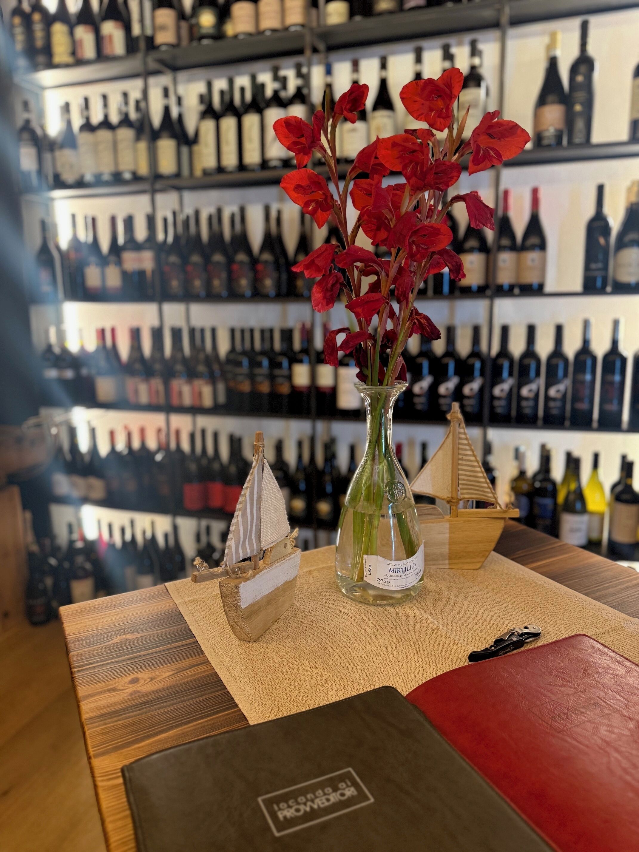 Una bottiglia di vino con etichetta, un modellino di nave in legno e un vaso con fiori rossi su un tavolo in un negozio di vinicoli.