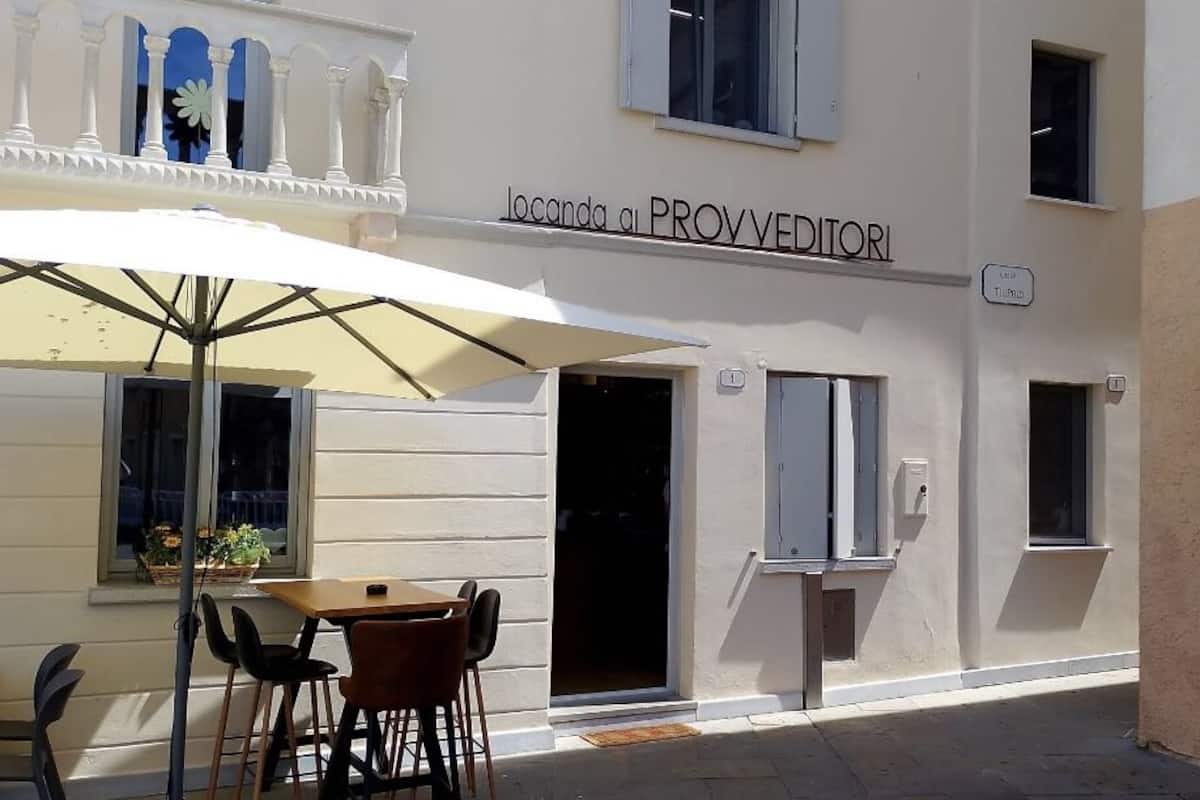 Un espace extérieur aménagé avec une table et des chaises sous un grand parasol, un bâtiment sur lequel on peut lire l’inscription « Locanda ai PROVEDITORI », et une fenêtre donnant sur la rue.
