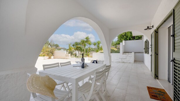 Un patio exterior blanco con una mesa y sillas, un sombrero tejido y vistas a palmeras y un edificio al fondo.