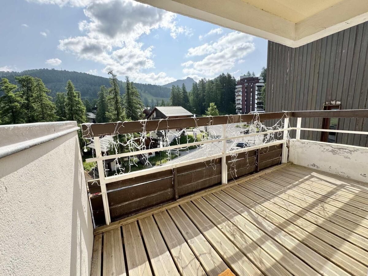Een balkon met een houten vloer en witte leuningen, met uitzicht op een bergachtig landschap met bomen en gebouwen.