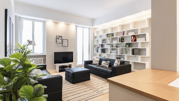 A modern living room with a sofa, ottoman, TV, bookshelf, and a large plant. - Easylife - Cosmopolitan Style in Porta Venezia (Milano)