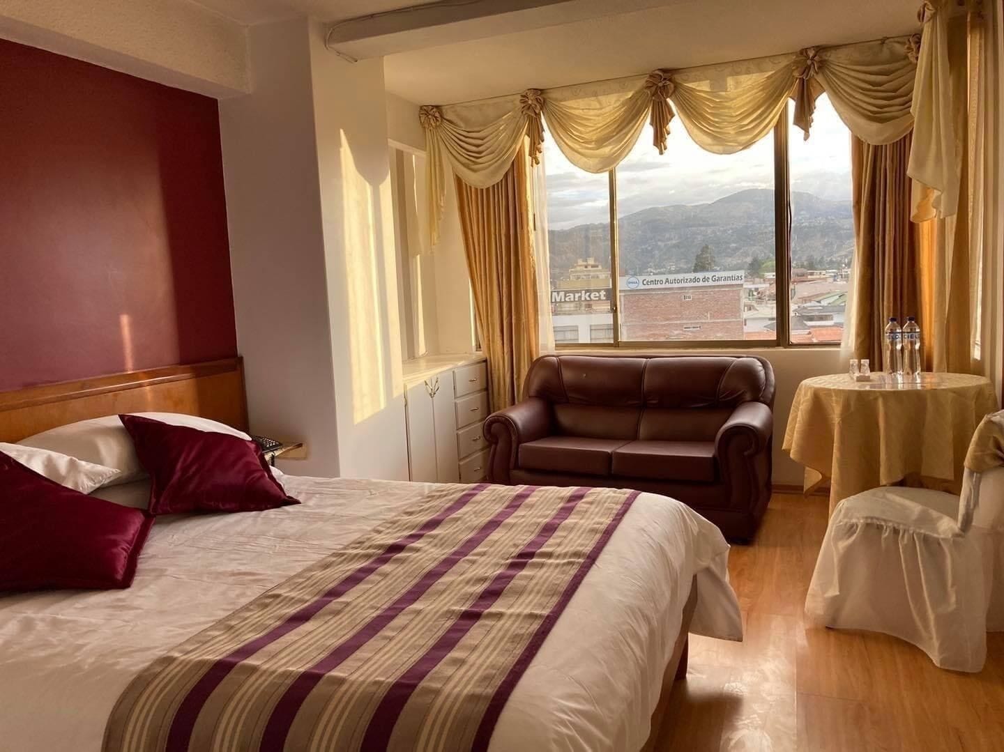 A hotel room with a large bed, a leather armchair, a small round table with a bottle, and a view of a cityscape through large windows.