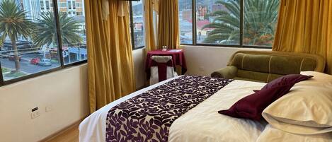 Habitación de hotel con cama, televisión montada en la pared, una mesita y vistas a palmeras y edificios.