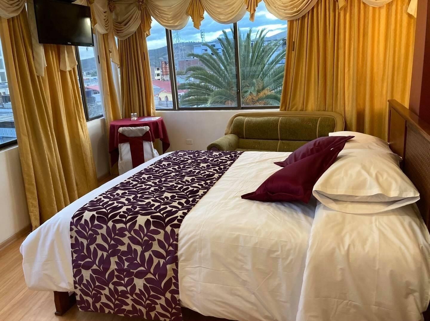 Une chambre d’hôtel avec un lit, un fauteuil vert, une petite table recouverte d’une nappe rouge, et une vue sur des palmiers et des immeubles, visible à travers de grandes fenêtres.