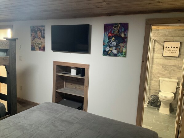 Ein Schlafzimmer mit einem Bett, einem an der Wand montierten Fernseher und einem kleinen Regal mit einer Fernbedienung.